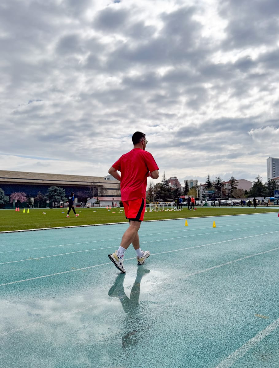 Yağmur sonrası ıslak atletizm pistinde tempolu yürüyüş yapan kırmızı tişörtlü katılımcı; arkada çim sahada ısınan diğer katılımcılar.